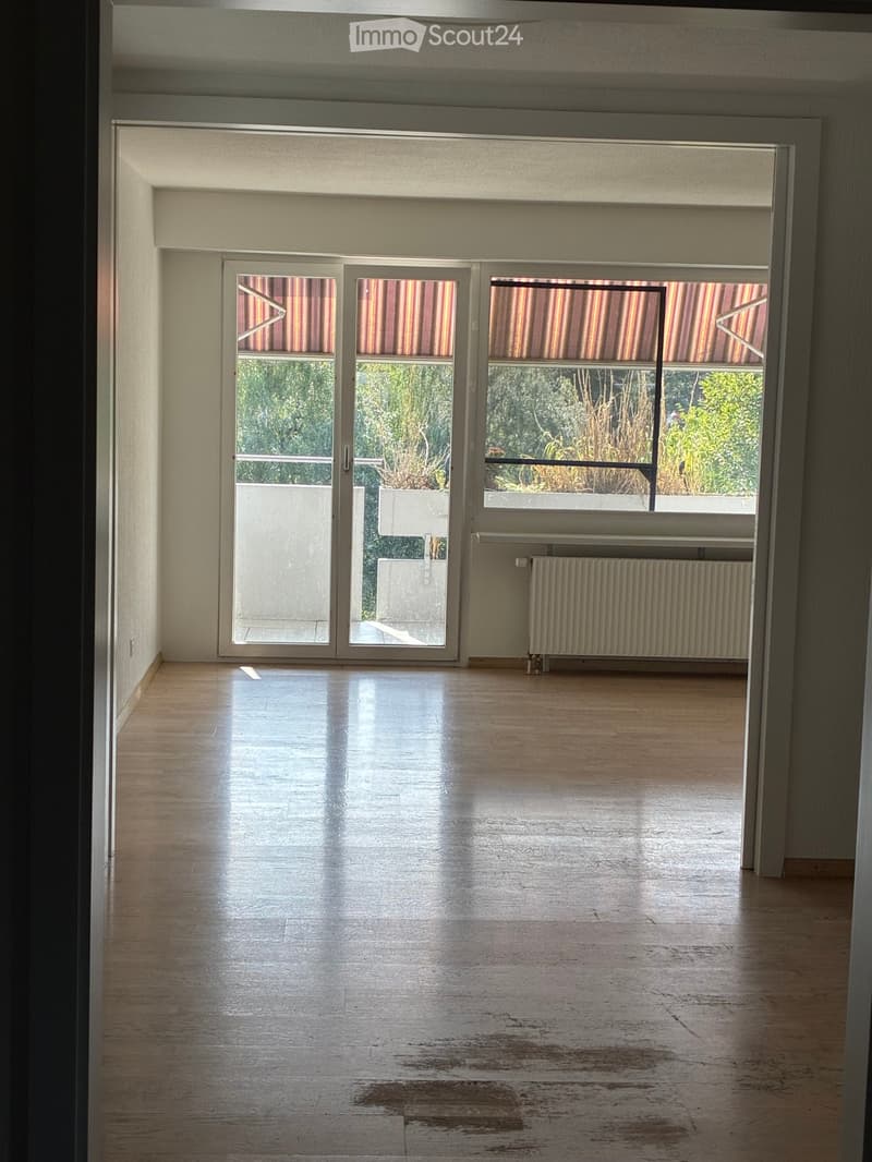 Empty room with wooden flooring, large sliding glass doors leading to a balcony, radiator, and visible outdoor greenery.
