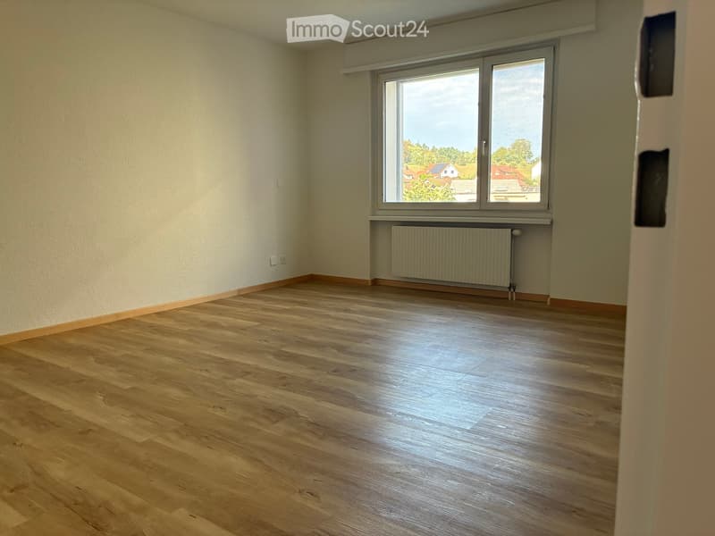 Empty room with wooden flooring, large window, radiator, and light-colored walls.
