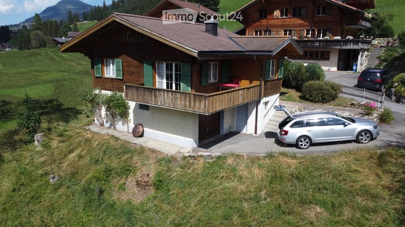 A two-story wooden house with green shutters, a balcony, and a garage. The house is surrounded by greenery and has a scenic mountain view in the background.