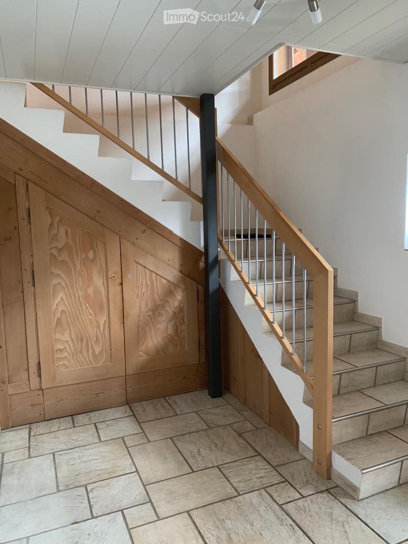 Modern staircase with wooden railings and metal balusters, leading to an upper level. The staircase features a sleek design with a central black column and warm wood tones.