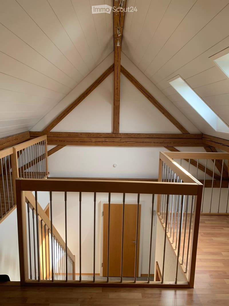 Spacious hallway with wooden floors, exposed wooden beams, and a modern staircase with metal railings. The ceiling features a sloped, vaulted design with skylights allowing natural light to enter.