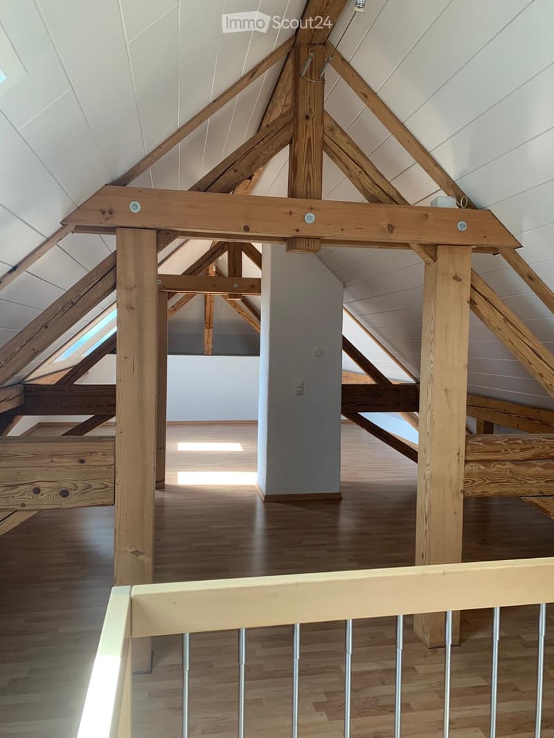 Open, airy attic space with exposed wooden beams and a wooden floor, featuring a railing and skylights allowing natural light to enter.
