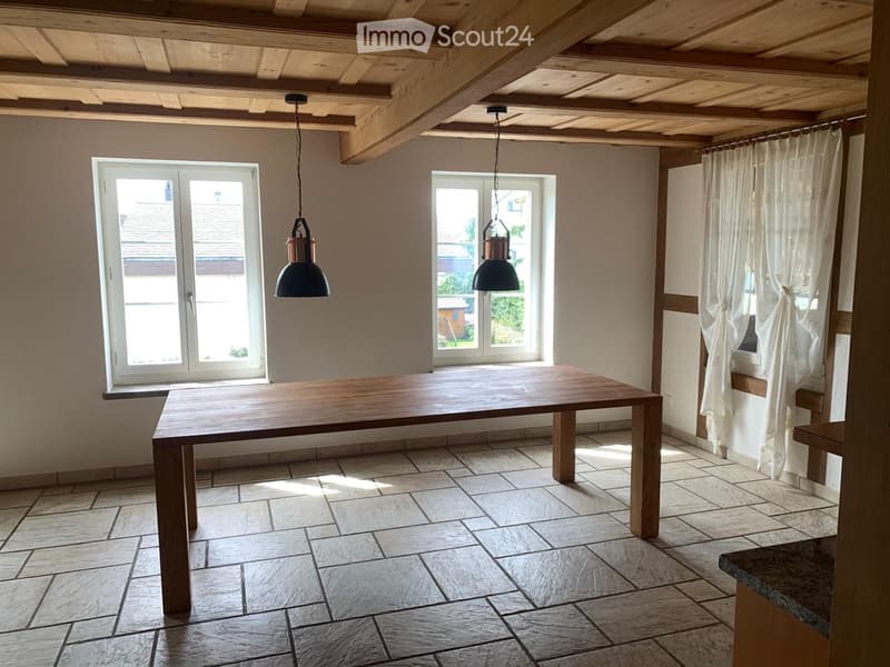 Bright, unfurnished room with exposed wooden ceiling beams, tiled floor, large windows with white curtains, and a wooden dining table.