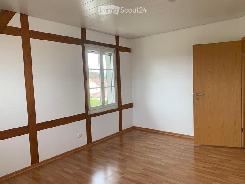 Empty room with wooden flooring, white walls, and a window with white frame. Wooden door on the right side.