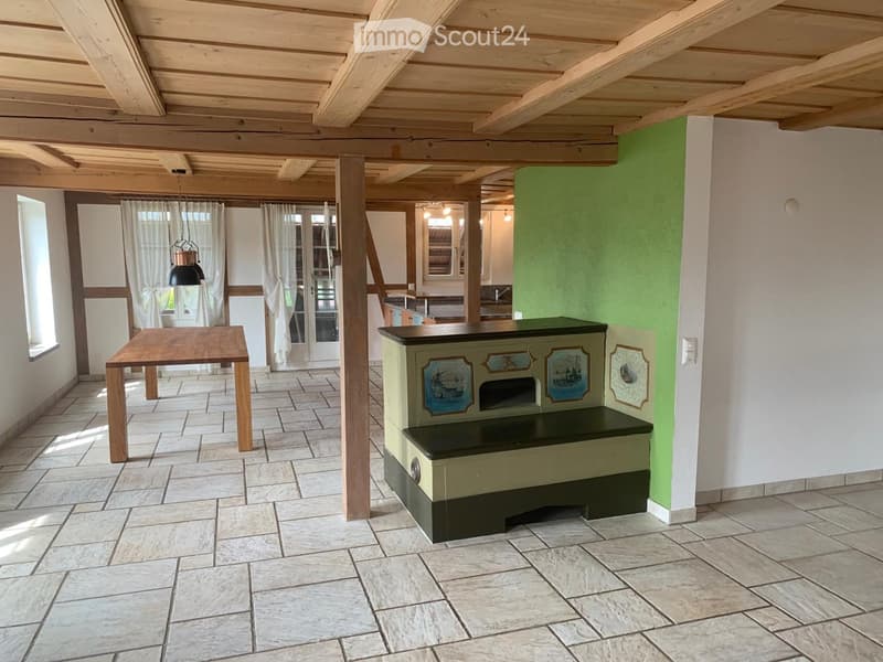 Bright, open room with exposed wooden beams, tiled floor, and large windows with white curtains. Features a wooden table, a painted bench with decorative panels, and a hanging light fixture.