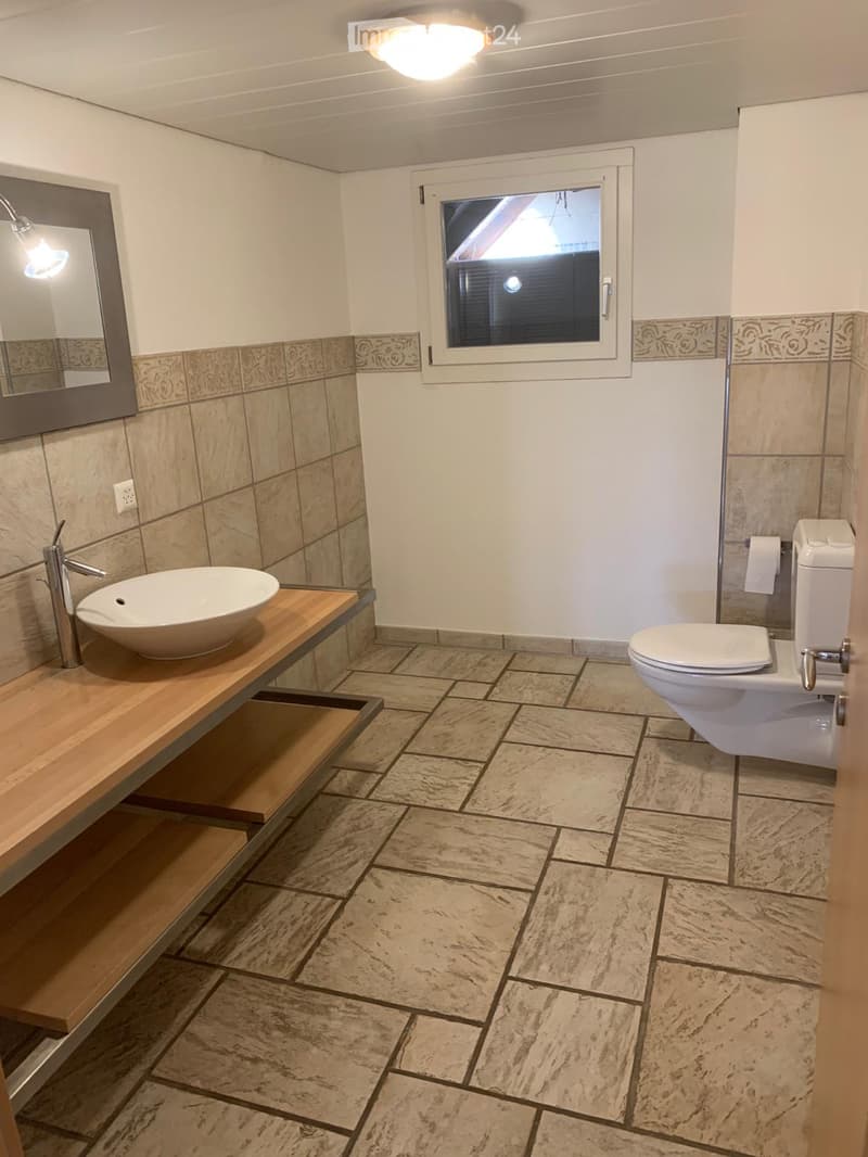 Modern bathroom with beige tiles, a wooden sink console, a white sink, a toilet, and a large mirror. Features a window with blinds and a ceiling light fixture.