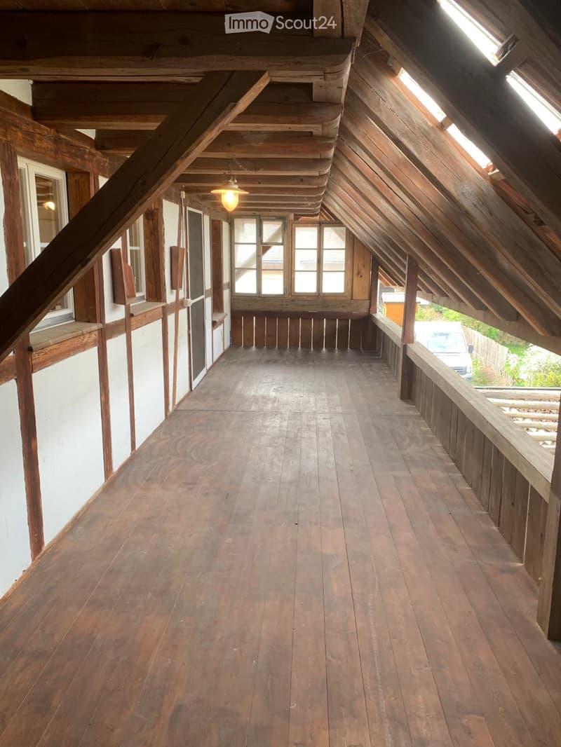 Wooden hallway with exposed beams, wooden floor, and windows on both sides, leading to an open area with a wooden railing.