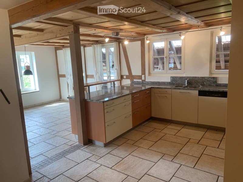 Bright, open kitchen with light-colored cabinets, granite countertops, and tiled flooring. Exposed wooden beams on the ceiling and large windows provide natural light.