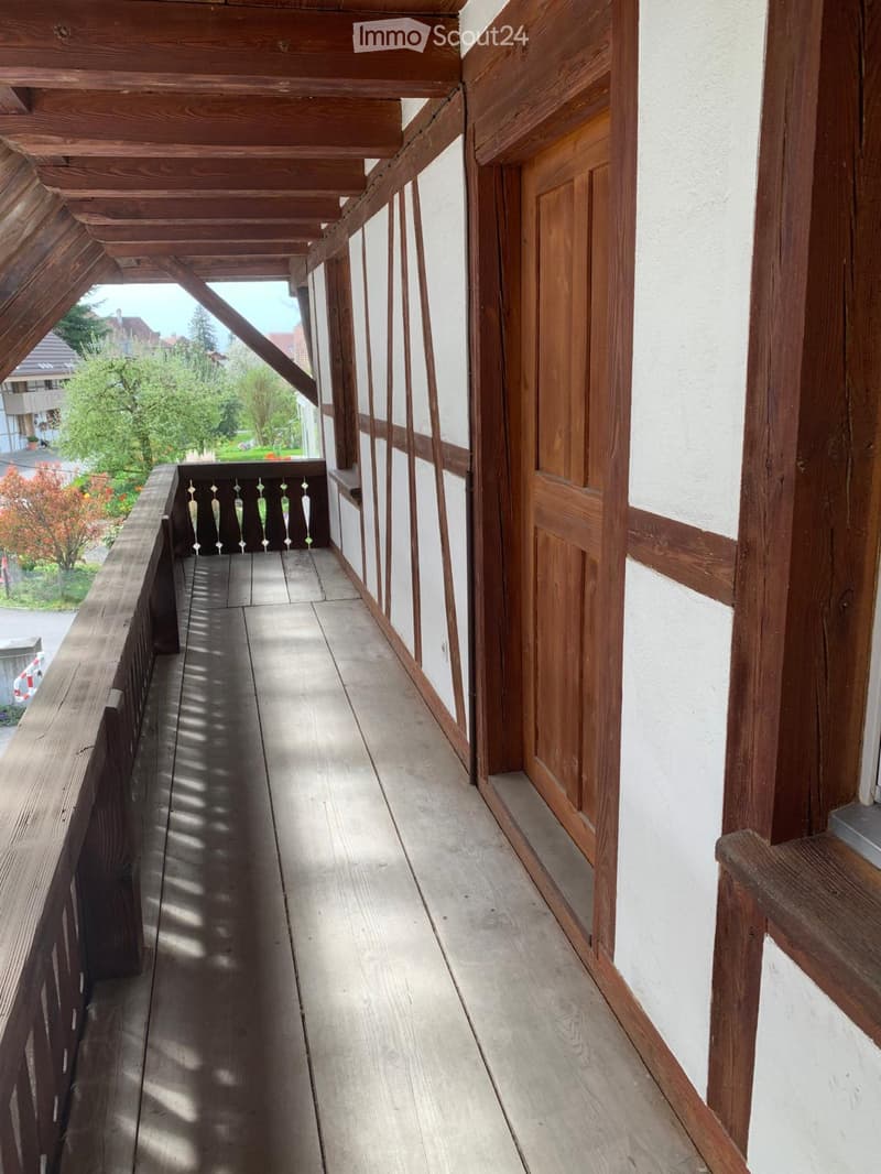 Wooden balcony with a railing, wooden floor, and white walls. The balcony has a door leading to the interior and overlooks a green area with trees.