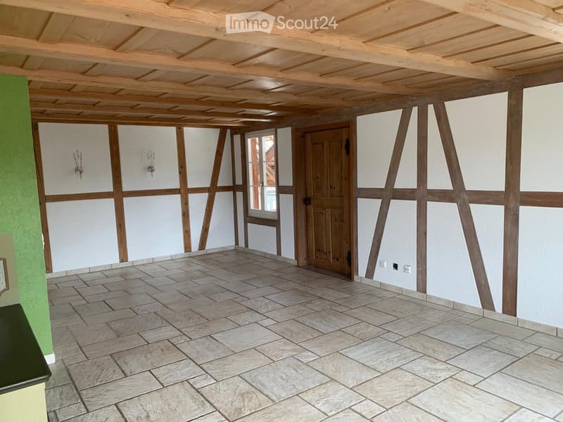 Empty room with exposed wooden beams, tiled floor, white walls, and a wooden door leading to another space.
