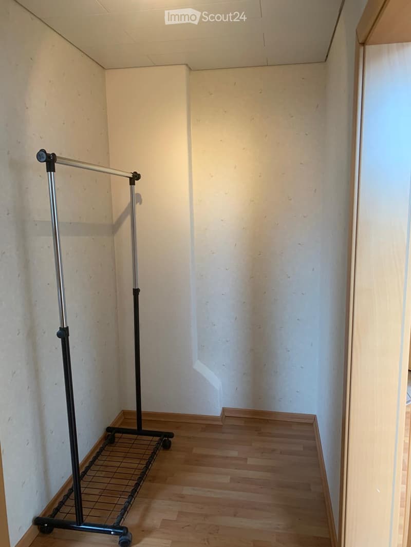 Empty room with wooden flooring and white walls, featuring a clothing rack with a grid shelf on wheels.