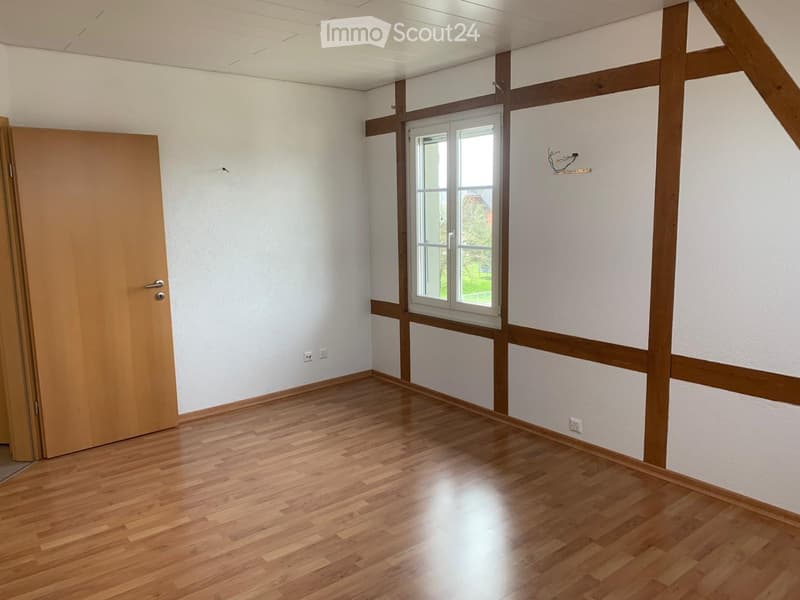 Empty room with wooden flooring, white walls, and a window with wooden trim. The room features exposed wooden beams and a partially open wooden door.