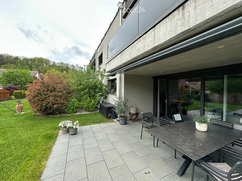 Modern patio area with a black dining table and chairs, surrounded by potted plants and a well-maintained lawn.