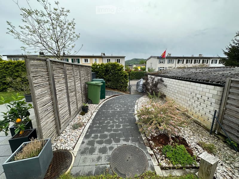 Garden area with paved pathway, wooden fence, potted plants, and green waste bins.