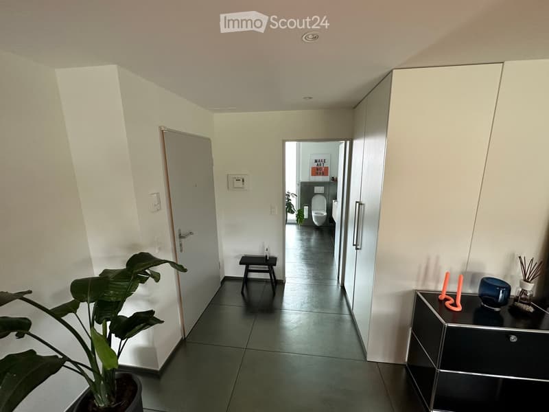 A modern hallway with white walls, dark tiled floor, and several doors leading to different rooms. A small black stool is placed near the center, and a potted plant is visible on the left side. A small table with decorative items is on the right side.