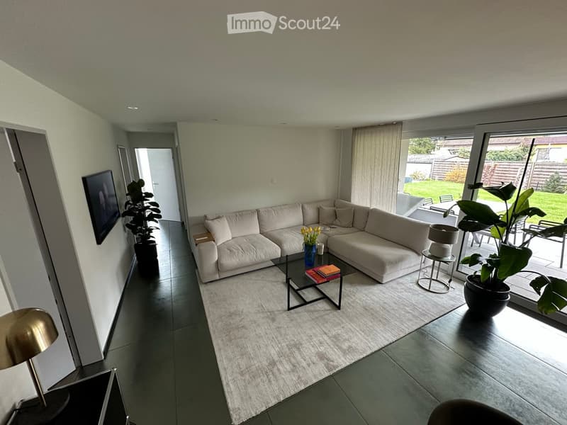 Modern living room with a large sectional sofa, glass coffee table, potted plants, and large windows leading to a patio.