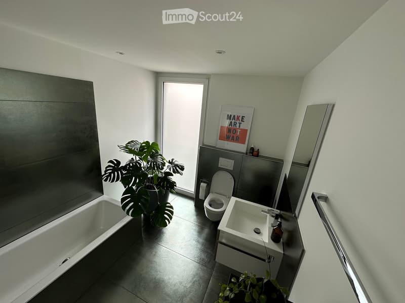 Modern bathroom with a bathtub, toilet, sink, and a large plant near the window. The space features sleek, contemporary design elements and a large mirror.