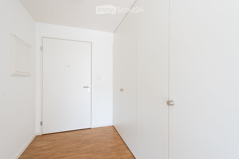Empty room with white walls and wooden flooring, featuring a closed white door and a small wall-mounted cabinet.