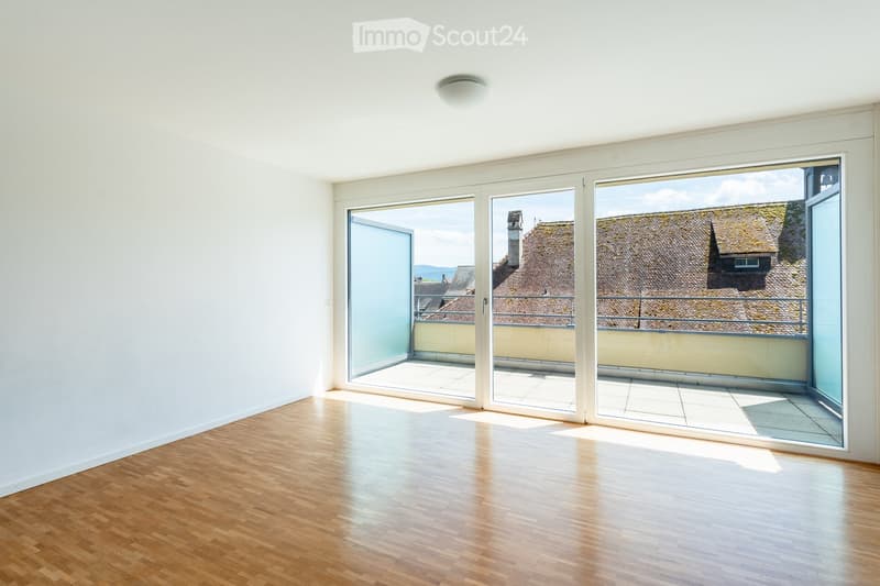 Empty room with wooden flooring, white walls, and large glass sliding doors leading to a balcony with a view of rooftops.