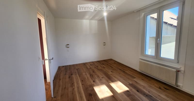 Empty room with wooden flooring, white walls, and large windows allowing natural light. Features a radiator under the window and a door leading to another space.