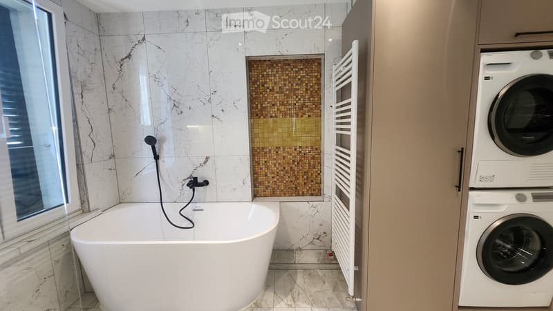 Modern bathroom with a white bathtub, marble tiles, a showerhead, a towel rack, and stacked washing/drying machines.