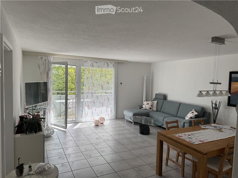 Bright living room with tiled floor, grey sectional sofa, coffee table, dining table with chairs, TV, floor lamp, and decorative items near the balcony door.