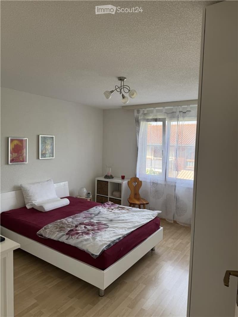Cozy bedroom with a white bed, red bedding, wooden floor, nightstand, and a small wooden chair near the window.