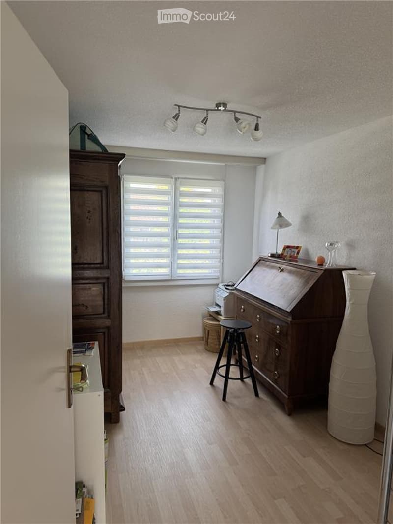 A well-lit study or home office with light wooden flooring, a large window with blinds, a wooden desk with a chair, a tall cabinet, and decorative items including a vase and picture frame on the desk.