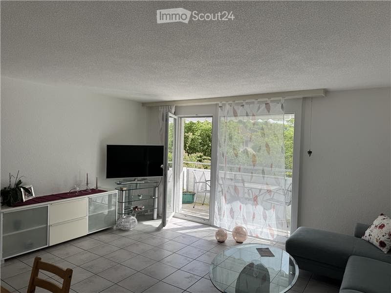 Modern living room with a grey sofa, glass coffee table, flat-screen TV on a white cabinet, and a sliding glass door leading to a balcony.