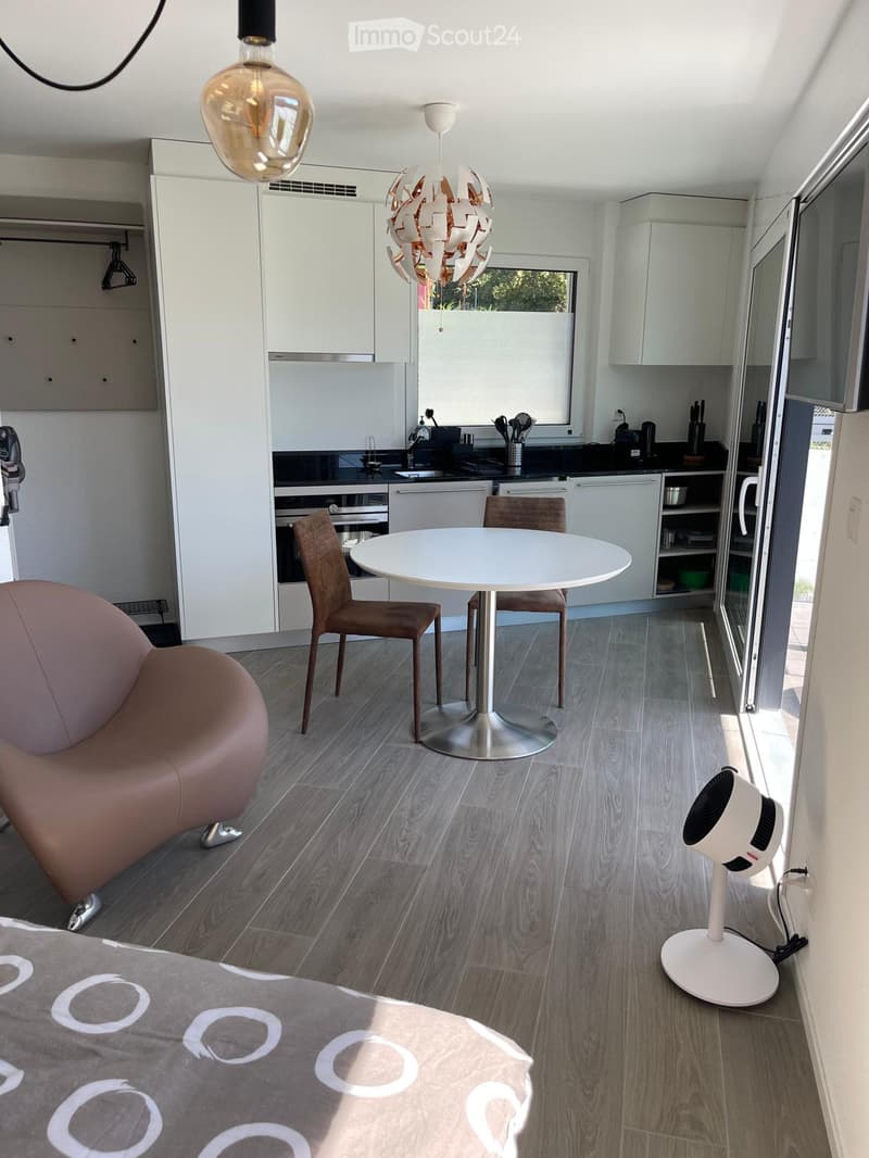 open plan kitchen with dining area, modern style, large dining table with 4 chairs, stainless steel kitchen island with sink, white cabinets, modern pendant light, wooden floor, open door, white sheet on bed