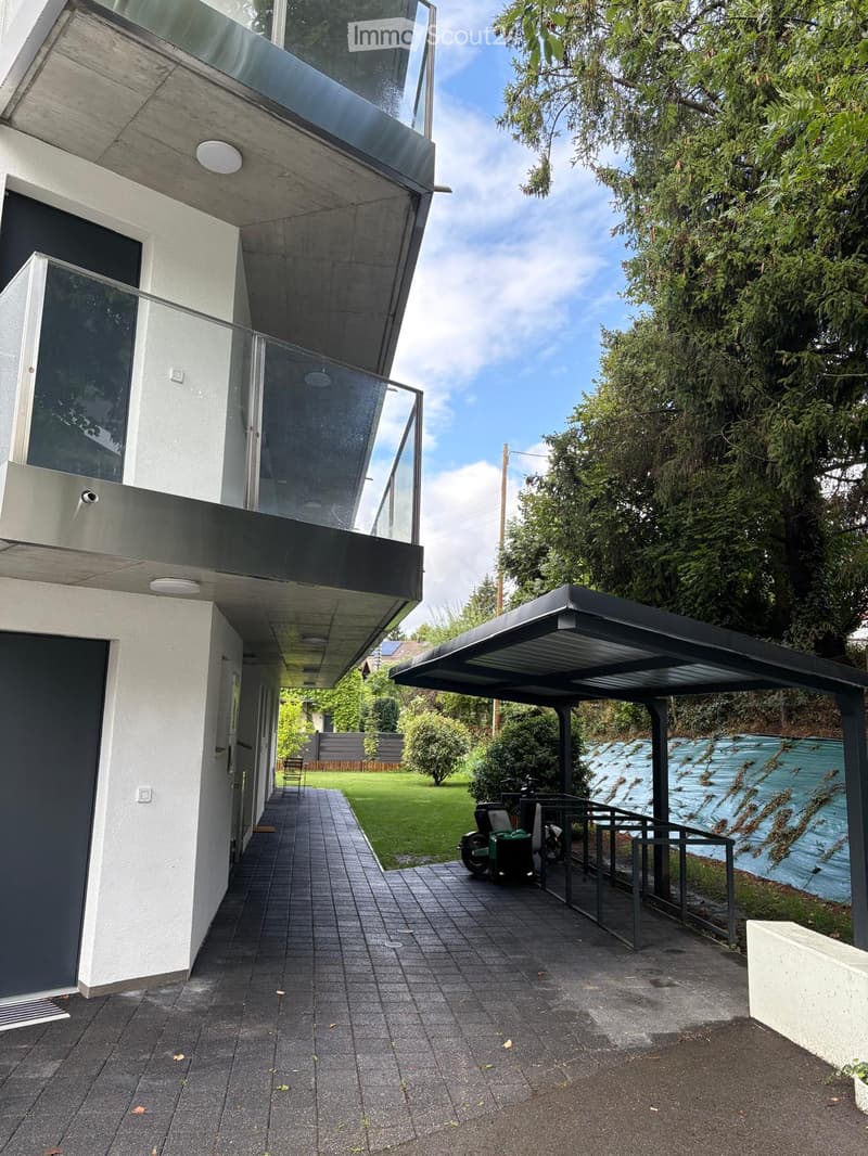 White modern house, gray balcony, metal carport, tiled pathway, green lawn, trees, clear sky