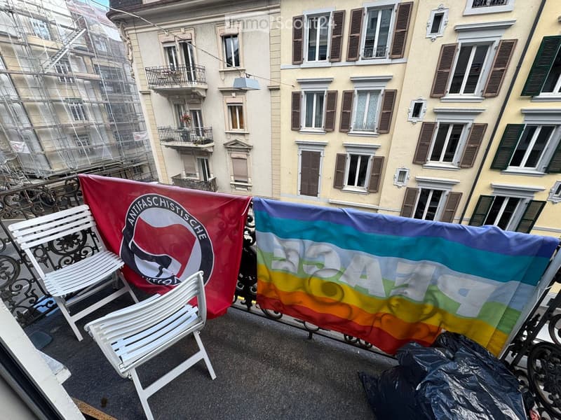 Balcony with two white chairs and two flags, one red and one rainbow-colored, set against a backdrop of multi-story residential buildings with balconies and shutters.