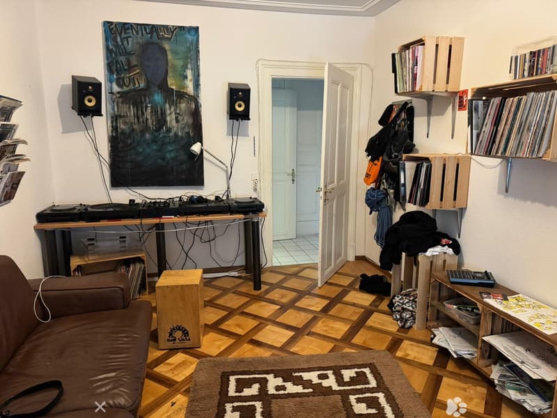 A cozy, well-furnished room with a wooden floor, a brown leather sofa, a wooden table with record players, shelves filled with books and records, a lamp, and a large poster on the wall.