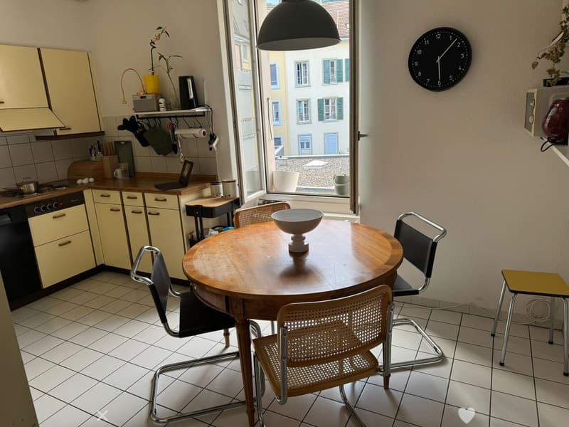 A compact kitchen with a round wooden dining table, four chairs, a dishwasher, oven, and cabinets. A window offers a view of the street, and a clock is mounted on the wall.