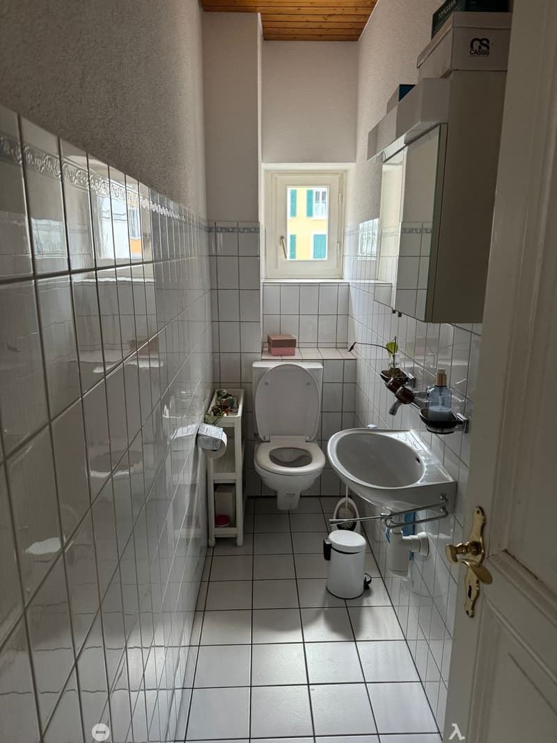 Small bathroom with white tiled walls and floor, featuring a toilet, sink, mirror, and cabinet. A window provides natural light.
