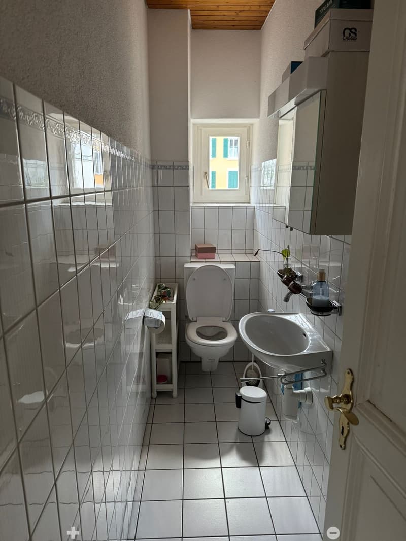 Small bathroom with white tiled walls and floor, featuring a toilet, sink, mirror, and cabinet. A window provides natural light.