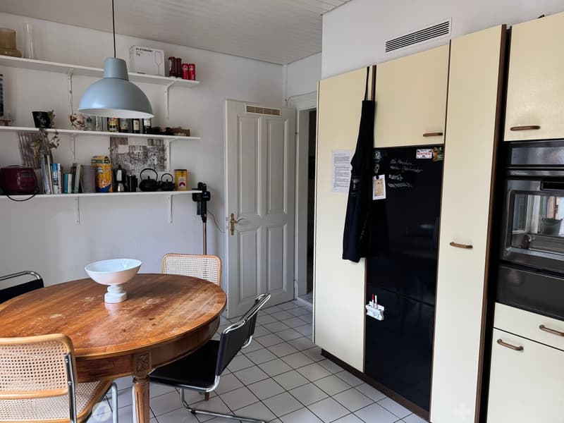 The kitchen features a round wooden dining table with a white bowl, surrounded by wicker and metal chairs. There is a refrigerator, oven, and built-in cabinets. Shelves hold various items including books, bottles, and a teapot.