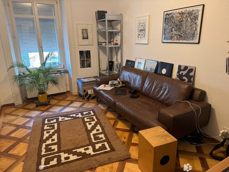 Modern living room with a brown leather sofa, wooden floor, large window with blinds, potted plant, shelf with books and decorative items, and artwork on the walls.