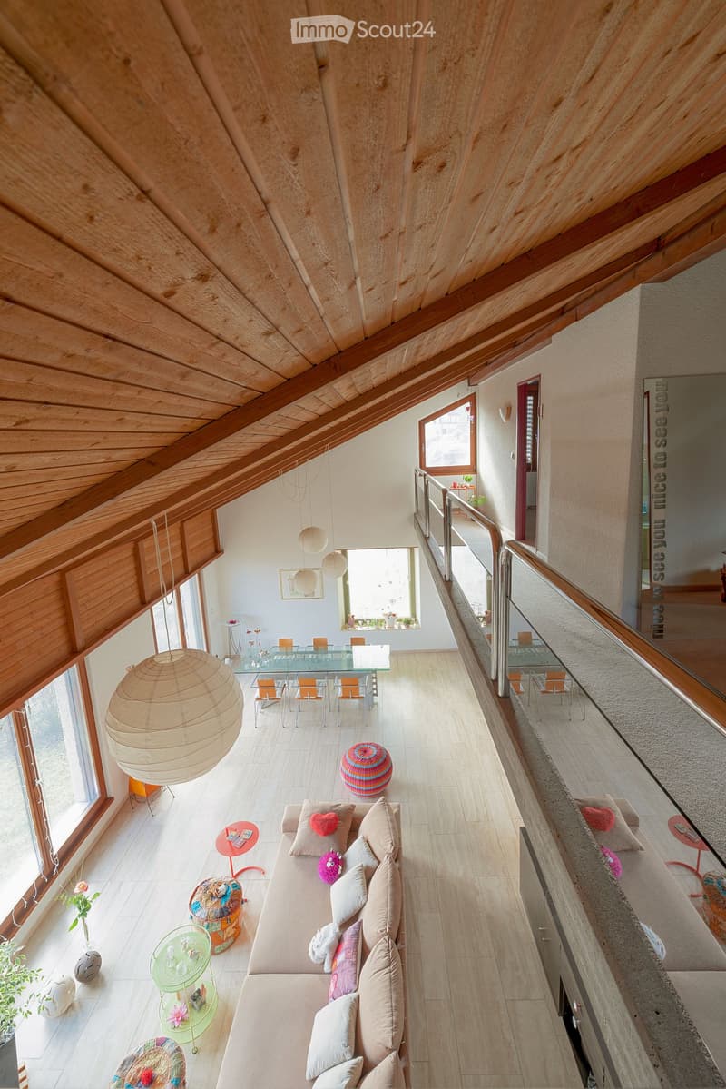 Open-plan living and dining area with a large sectional sofa, colorful cushions, a glass dining table with chairs, and a hanging paper lantern. The space features wooden beams on the ceiling and large windows letting in natural light.