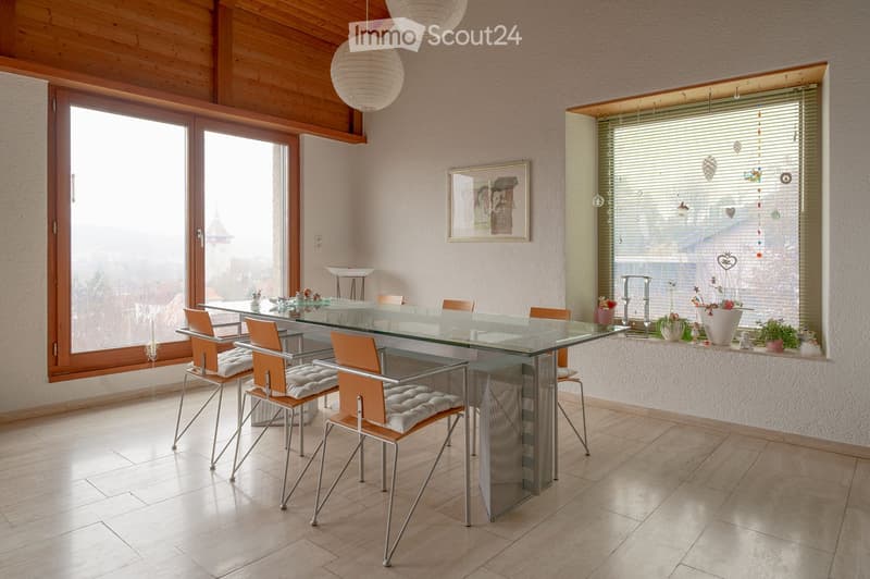 Dining room with a glass table, six chairs, a window with blinds, and a view of the outside. There are decorative items on the windowsill and a framed picture on the wall.