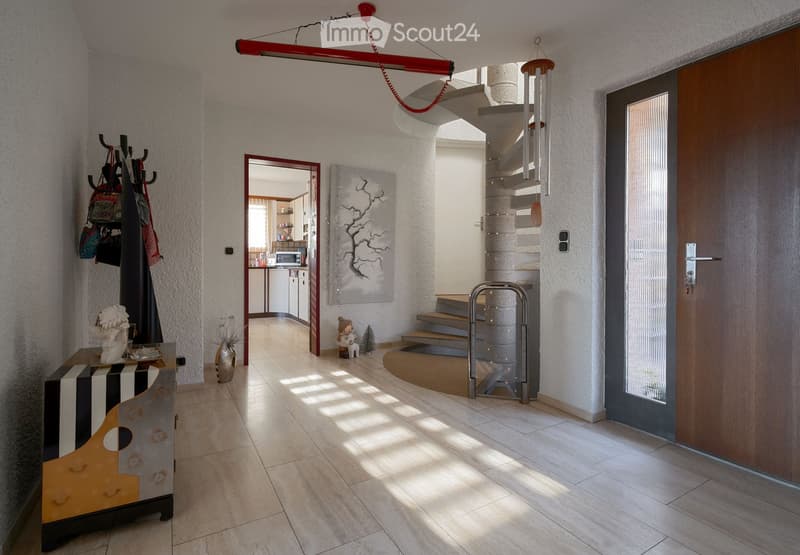 Bright hallway with tiled flooring, a staircase leading upwards, a coat rack with bags, a small table with a decorative item, and a doorway to a kitchen area.