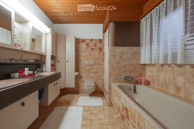 Bathroom with a double sink, mirror, toilet, bathtub, and floral-patterned tiles. Wooden ceiling and white curtains on the window.