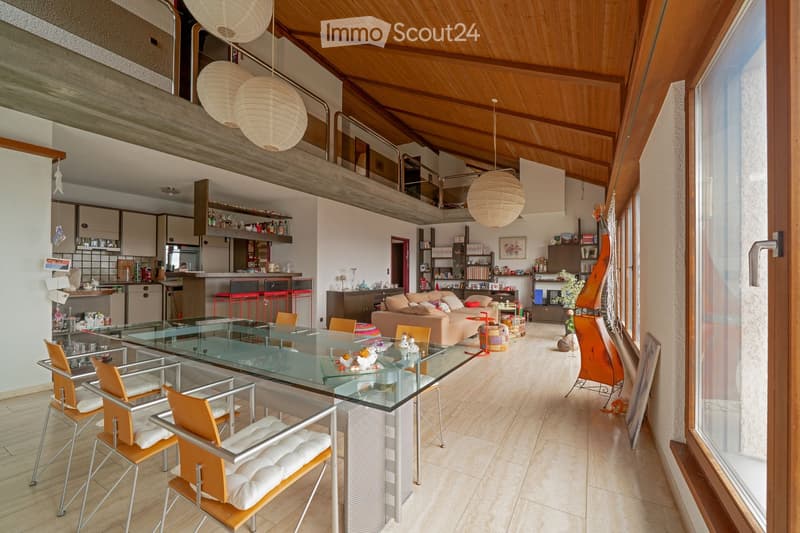 Open-plan living and dining area with a glass dining table, modern chairs, a sofa, kitchen area with counter and stools, and decorative items including a guitar and a flower vase.
