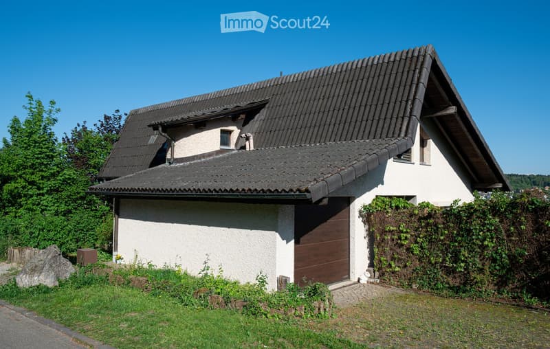 Two-story house with a dark tiled roof, white walls, and a wooden garage door. The house is surrounded by greenery and has a small garden area.
