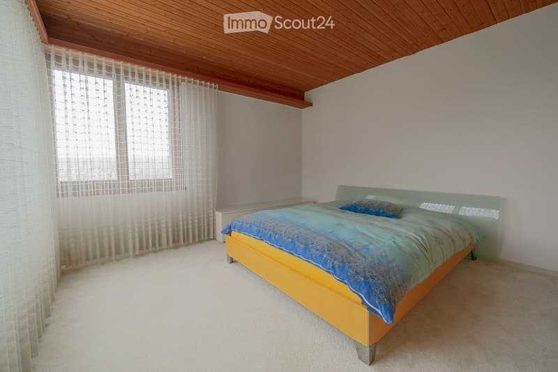 Bedroom with a wooden slatted ceiling, a large window with sheer curtains, a bed with a blue and green duvet, and a matching nightstand.