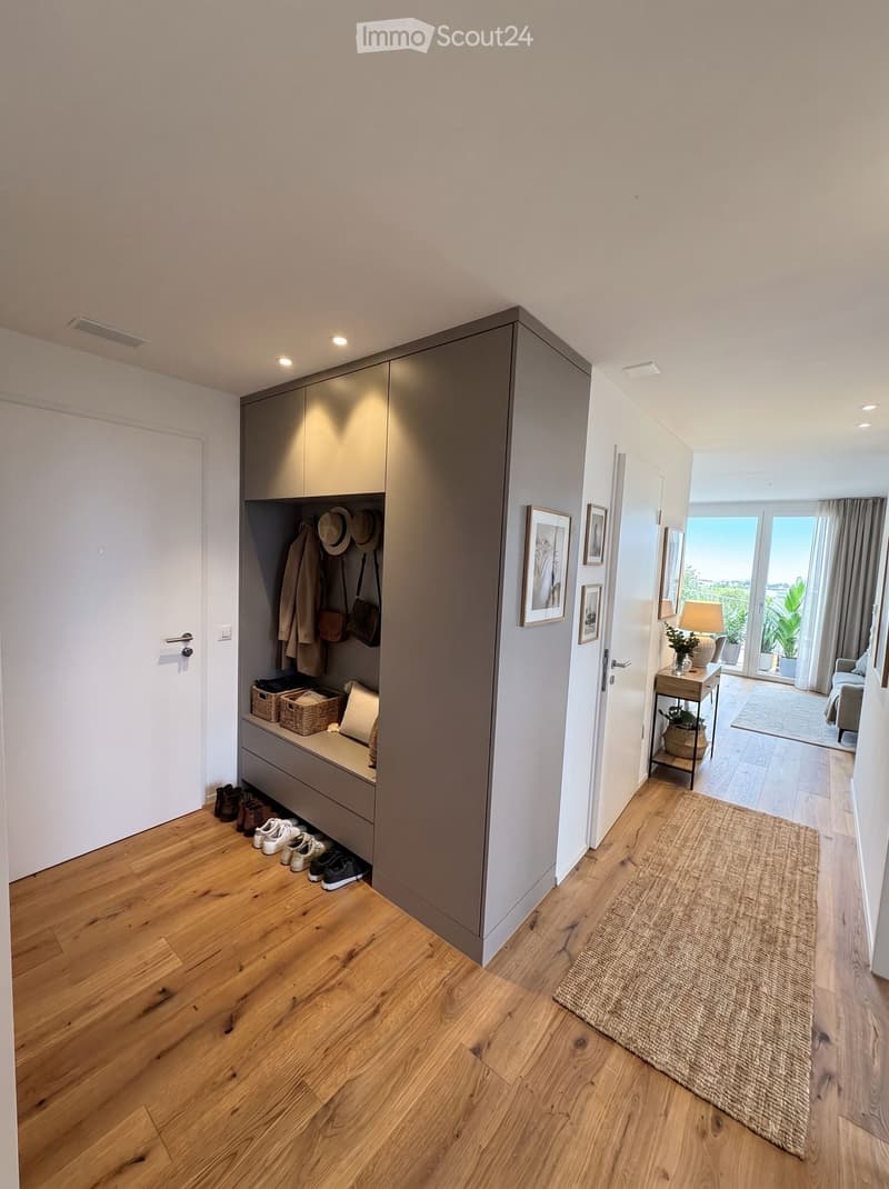 Modern hallway with wooden flooring, built-in storage unit, shoes, coats, and bags. A small table with a lamp and plant is visible, leading to a living area with a sofa and large windows.