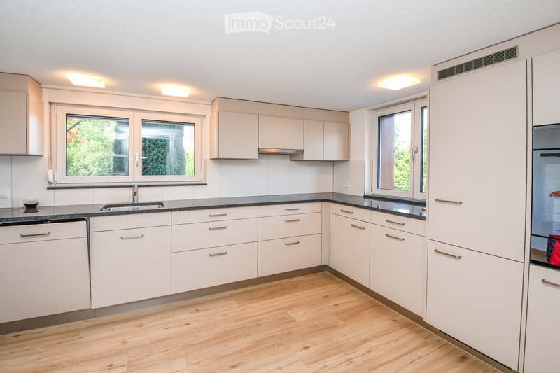Modern kitchen with light-colored cabinets, wooden flooring, and stainless steel appliances including a sink, dishwasher, and refrigerator. Large windows provide natural light.