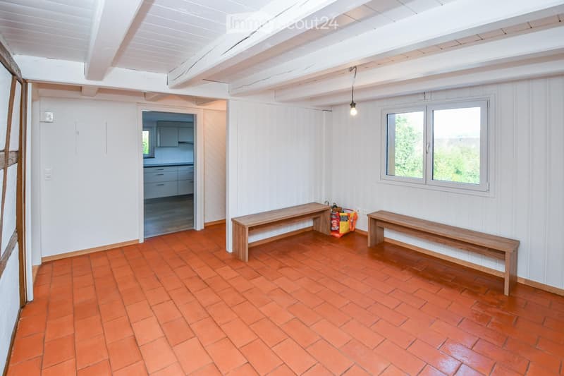 A bright, unfurnished room with white wooden panel walls, terracotta tiled floor, and two wooden benches placed against the wall. A doorway leads to another room, and a window allows natural light to enter.