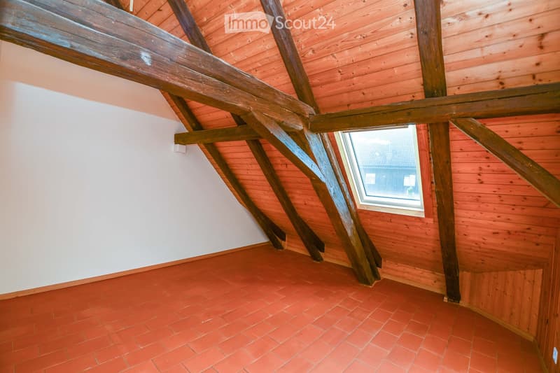 Empty attic space with wooden beams, red brick floor, and a small window showing a view of the roof.