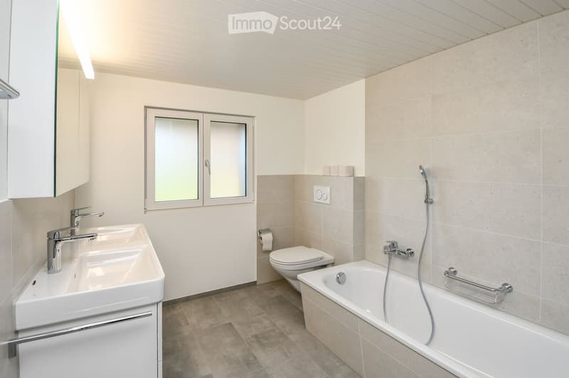 Modern bathroom with a white sink, toilet, bathtub, and a large mirror. The room features beige tiles and a window with frosted glass.